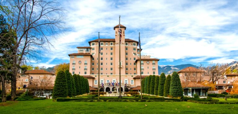 Grand hotel building with central tower, surrounded by trees and gardens under a partly cloudy sky.