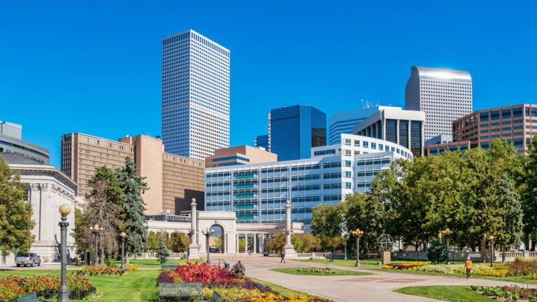 City skyline with modern and older buildings viewed from a landscaped park featuring flower beds and trees under a clear blue sky.