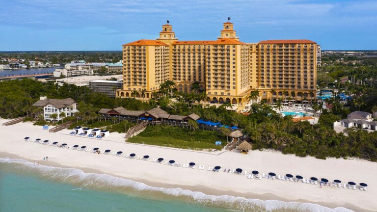 Beachfront resort with multiple buildings and orange roofs, set against a backdrop of trees and a blue sky. The sandy beach has rows of lounge chairs and umbrellas near the shoreline.