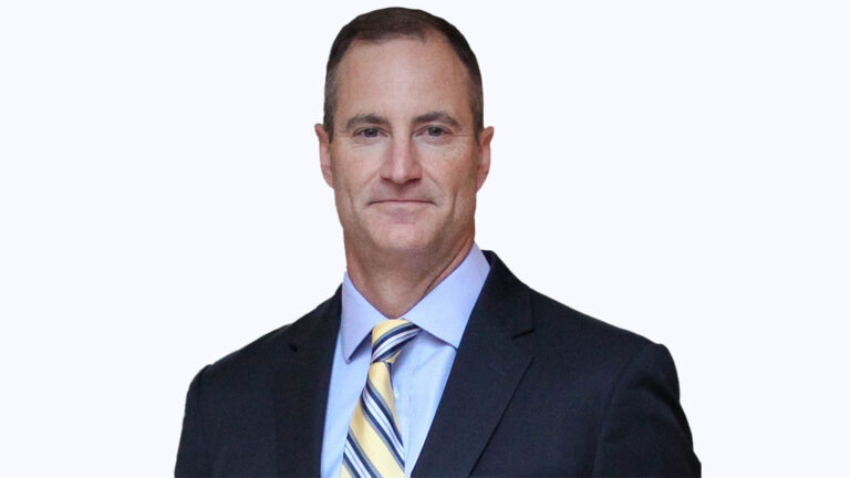 A man in a suit and tie smiles at the camera against a plain background.