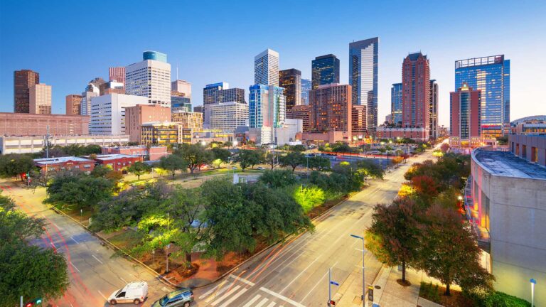 Aerial view of a cityscape with tall skyscrapers, green spaces, and roads. The scene is lit with a mix of natural and artificial light, indicating early evening or morning.