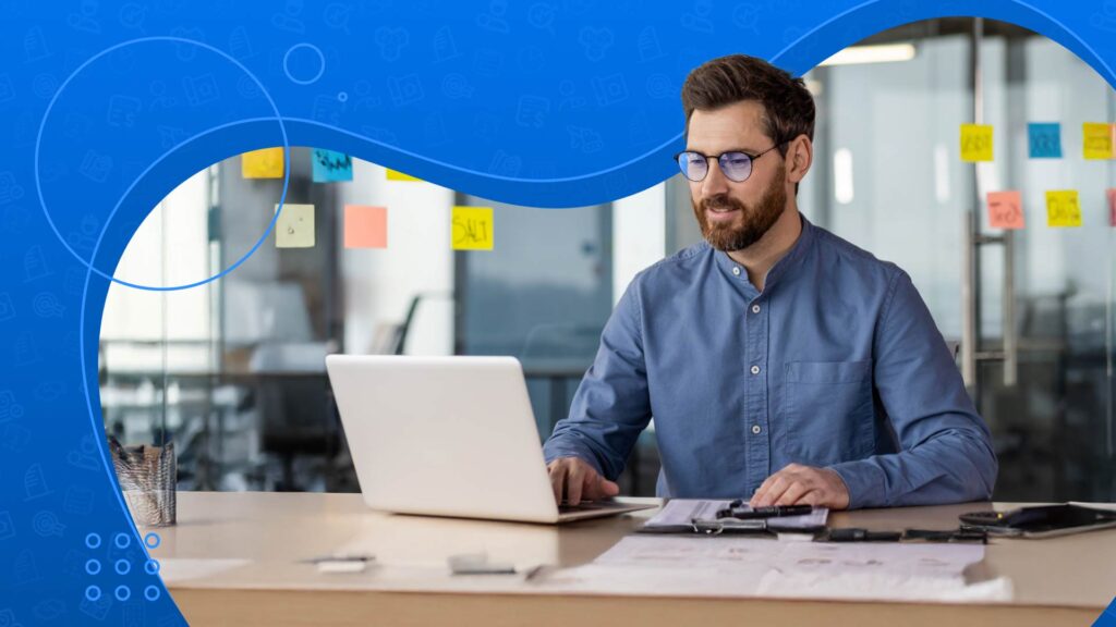 A man in glasses works on a laptop at a desk with sticky notes on a glass wall in the background.