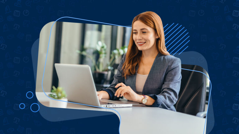 Woman in a blazer smiling while using a laptop at a desk in an office setting.
