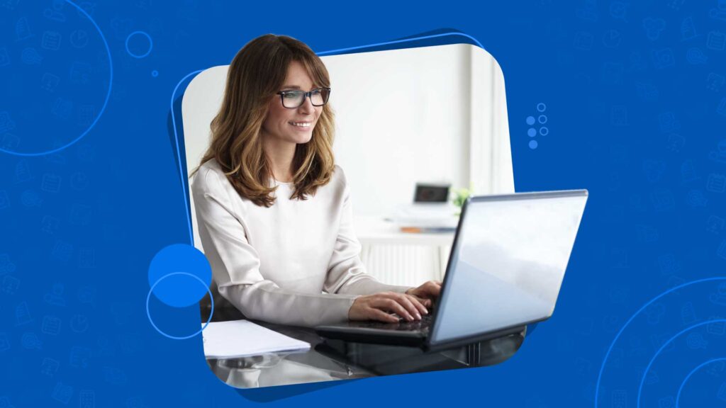 A woman with glasses, smiling, types on a laptop in a bright office space.