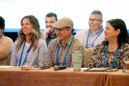 powerup25-what-to-expect-01 A group of people sitting at a table, smiling, and facing forward. They are wearing conference lanyards.