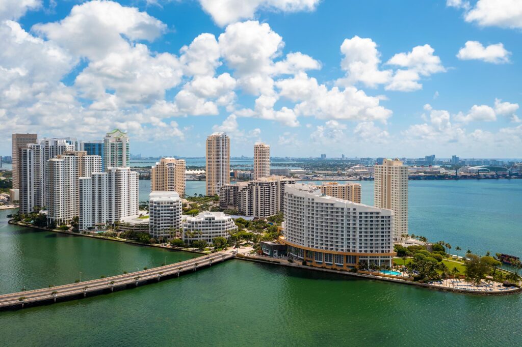 Aerial view of the coastal city, Miami, with tall buildings near the waterfront, a bridge connecting the area, and a cloudy sky above.