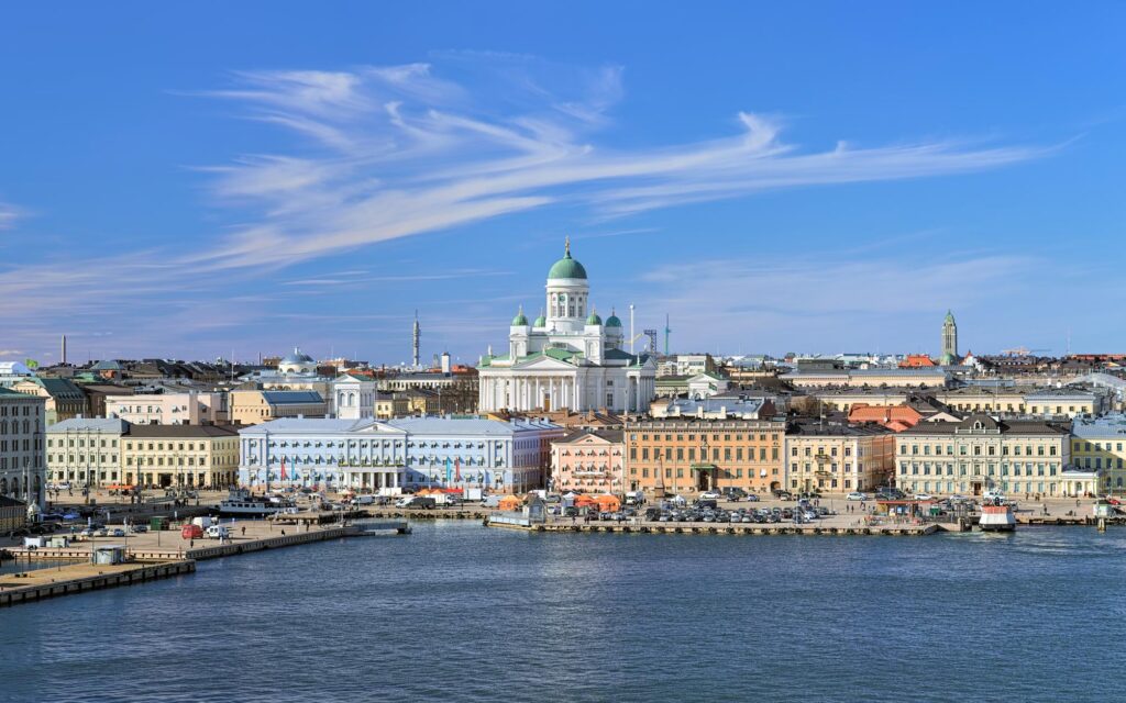 Skyline of Helsinki with the white Helsinki Cathedral at the center, surrounded by colorful buildings, and a body of water in the foreground under a blue sky.