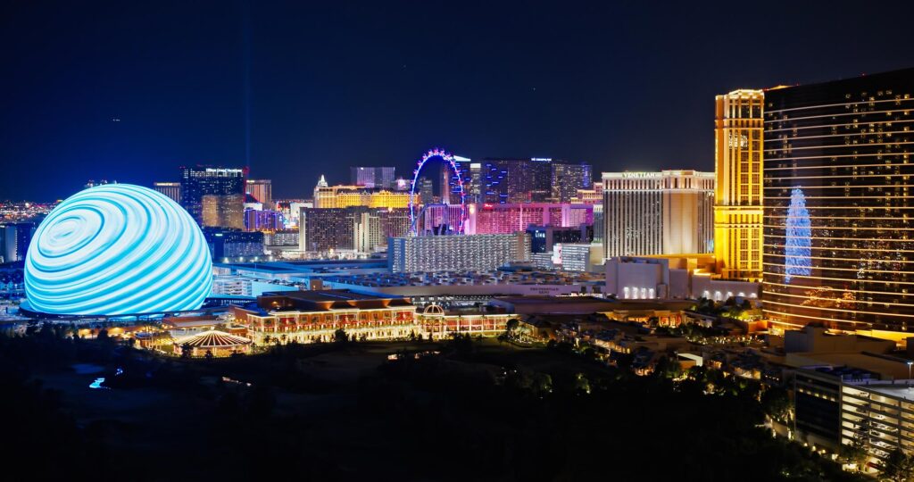 Night view of Las Vegas with brightly lit spherical building and Ferris wheel, surrounded by illuminated skyscrapers and city lights.