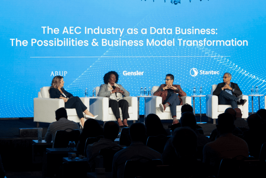 Four panelists sit on stage discussing the AEC industry as a data business, with a presentation screen displaying the event topic behind them.