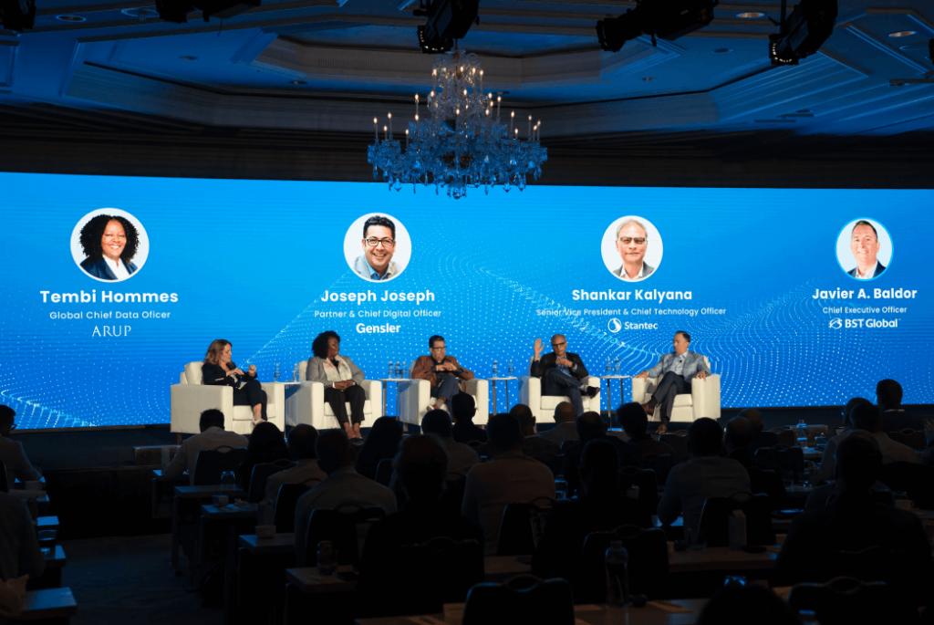 Five panelists sit on a stage in front of a large digital screen displaying their names, titles, and photos, speaking to an audience in a conference setting.