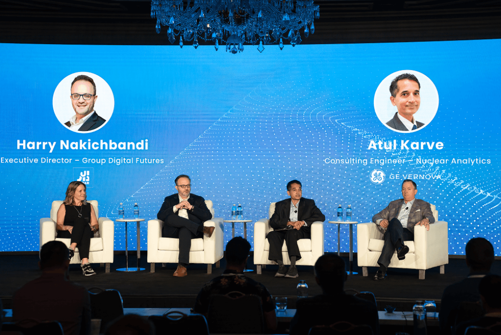 Four panelists sit on stage in front of a blue screen displaying names and titles: Harry Nakichbandi and Atul Karve. Audience members are visible in the foreground.