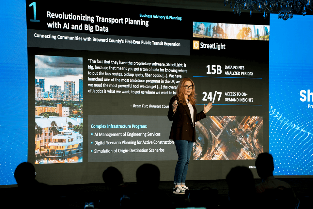 Shannon Miller stands on stage presenting a slide about AI and big data in transport planning, highlighting data points, 24/7 insights, and engineering service management.