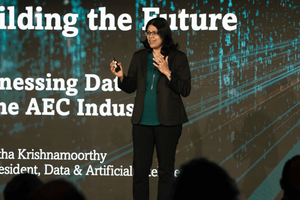 A woman stands on stage giving a presentation about data and artificial intelligence in the AEC industry, with a projected screen displaying her topic and name behind her.