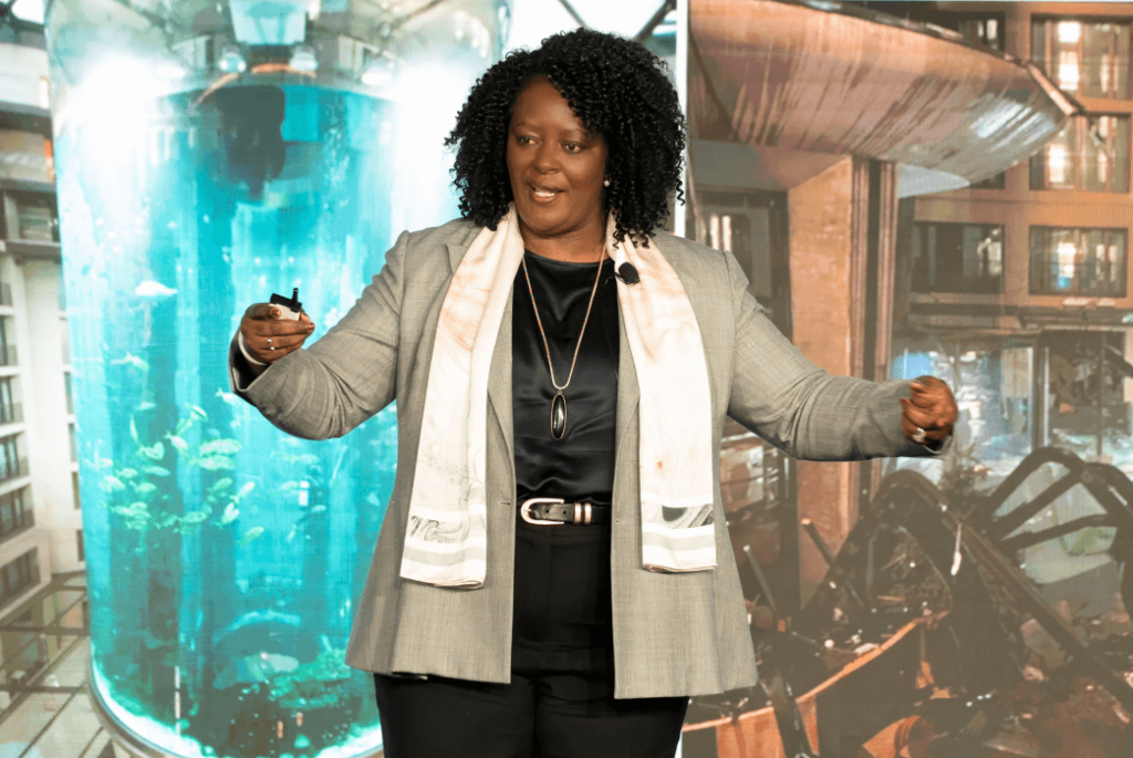 Tembi Hommes in a gray suit speaks on stage with a microphone headset, gesturing with both hands; a large aquarium and industrial setting are in the background.