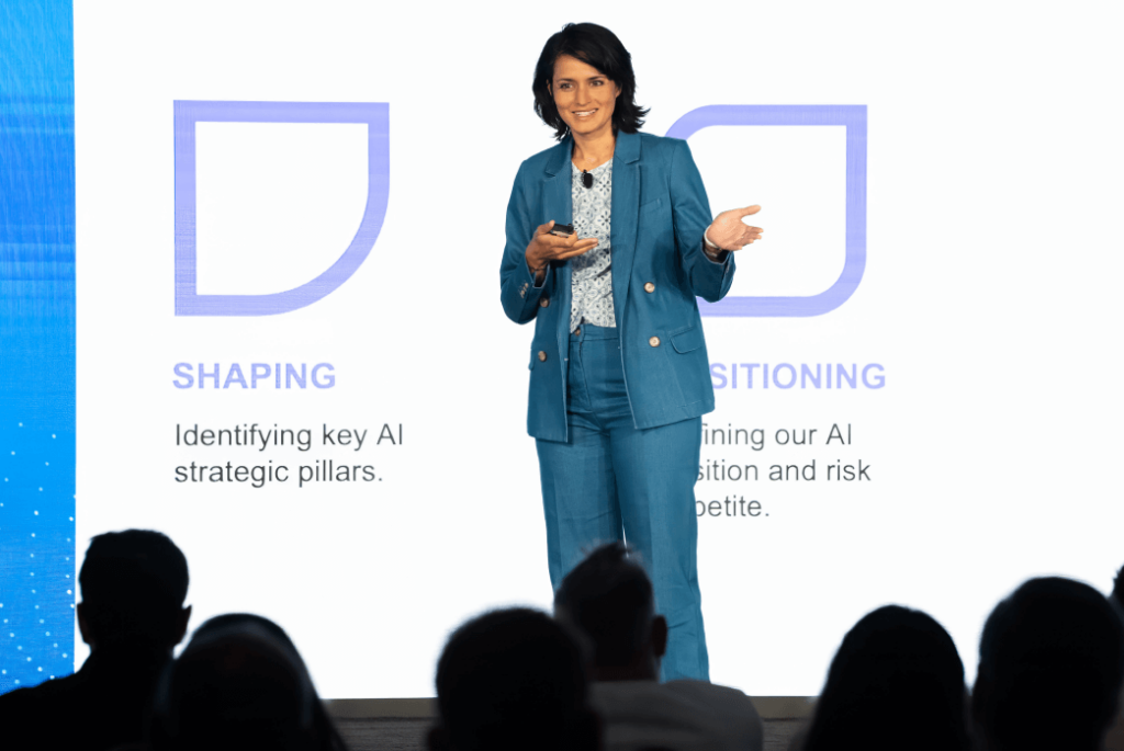 A woman in a blue suit speaks on stage in front of a screen displaying AI strategy concepts to an audience.