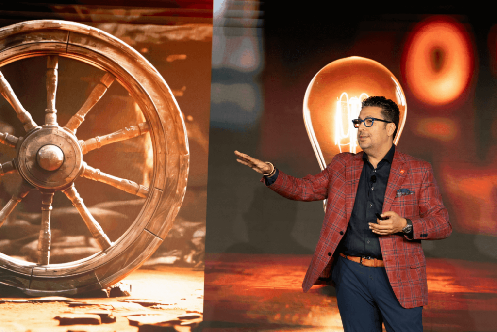 A man in a red checked blazer gives a presentation, gesturing toward a screen displaying an image of a wagon wheel and a light bulb.