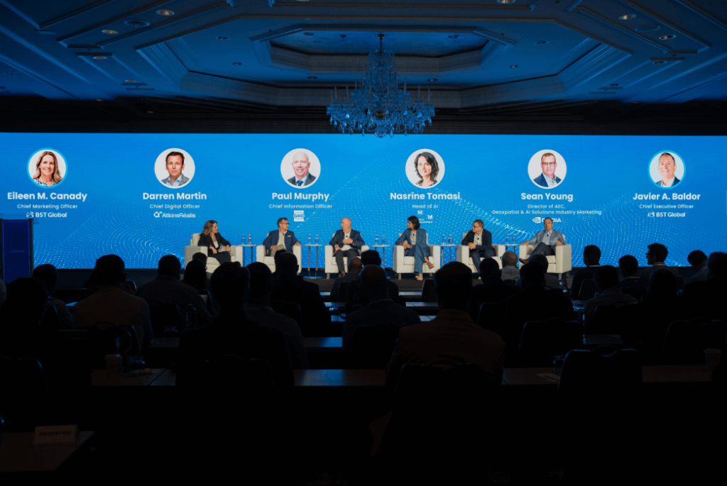 A panel of six speakers sits on stage in front of a large blue screen displaying their names and titles during a conference, with an audience seated in the foreground.