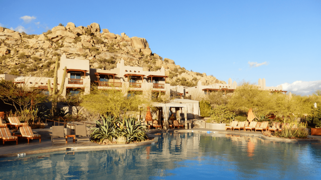 A serene outdoor pool area with lounge chairs is surrounded by desert landscape and rocky hills under a clear blue sky. Desert-style buildings are visible in the background.