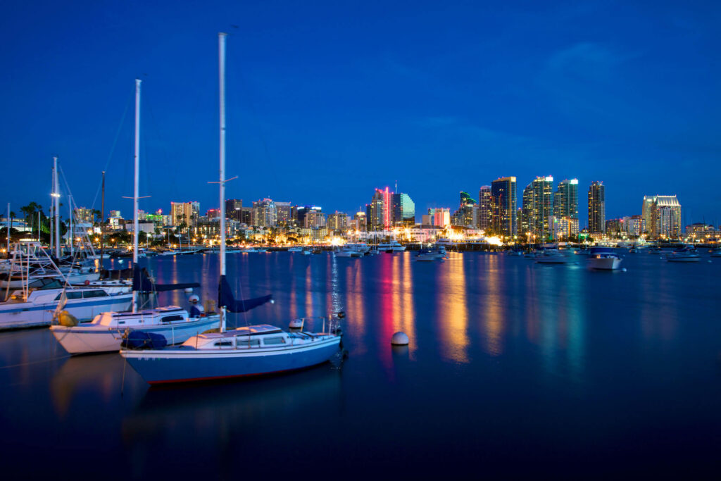 Sailboats are docked in a marina at dusk with city skyscrapers in the background and colorful reflections on the water.