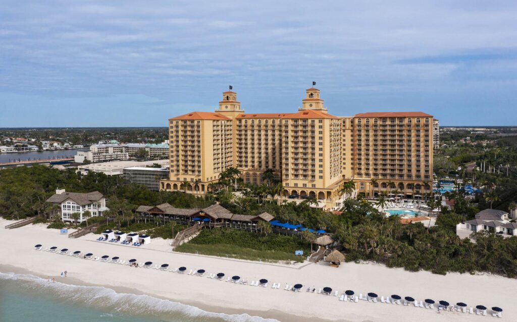 A large beachfront resort with two central towers, multiple terraces, pools, and beach chairs lined up along a white sandy shoreline under a partly cloudy sky.