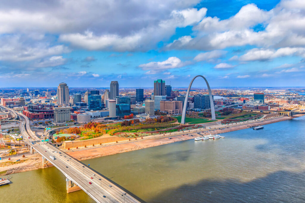 Aerial view of downtown St. Louis, Missouri, featuring the Gateway Arch, city buildings, a bridge, and the Mississippi River under a partly cloudy sky.