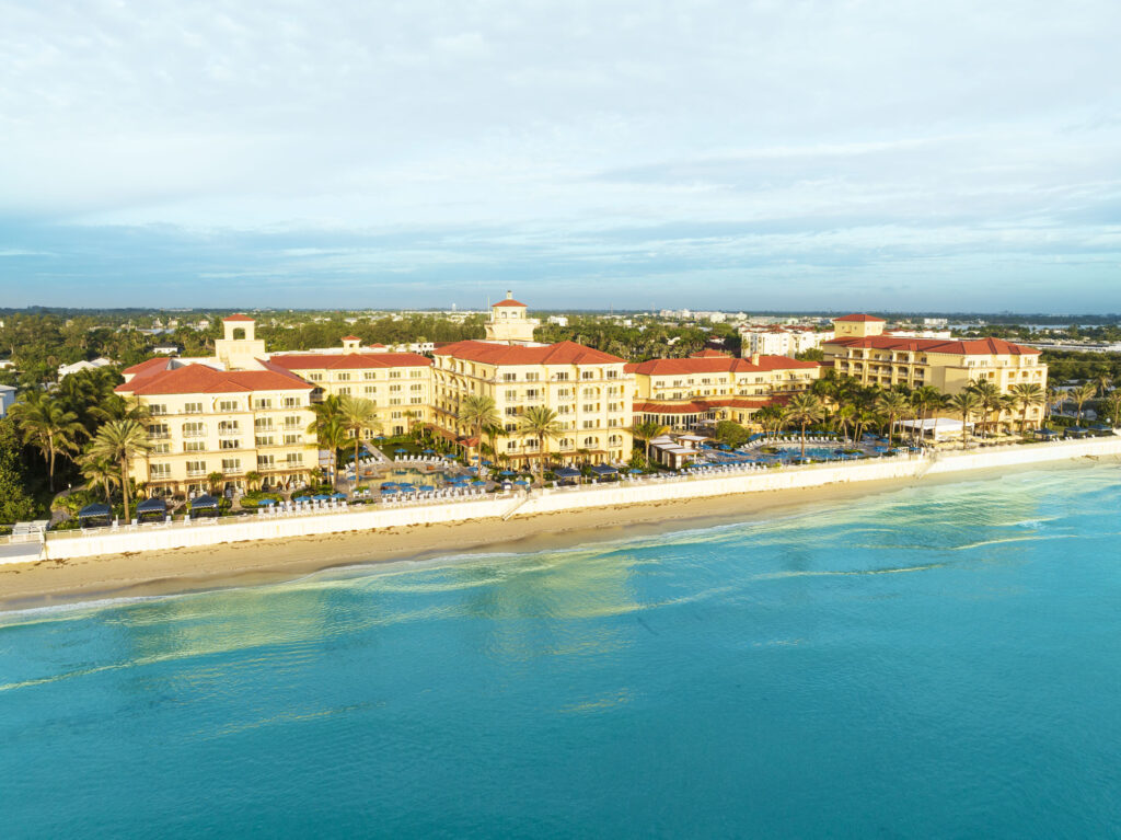 A large beachfront hotel with red-tiled roofs and yellow walls sits along a sandy shoreline, facing calm turquoise water under a partly cloudy sky.