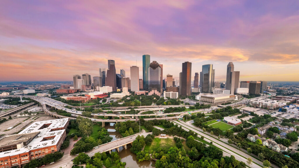 Aerial view of downtown Houston, Texas, at sunset, showing skyscrapers, highways, green spaces, and surrounding neighborhoods.