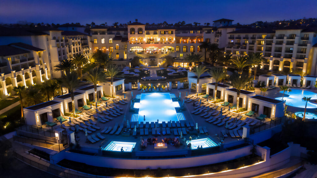 Aerial view of a luxury resort at night, featuring multiple pools, palm trees, lounge chairs, and illuminated buildings.