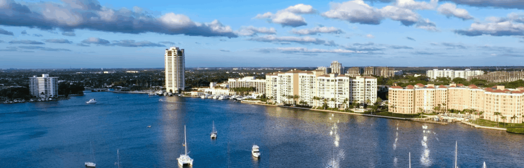 High-rise and mid-rise waterfront buildings line a calm bay with several anchored boats under a blue sky with scattered clouds.