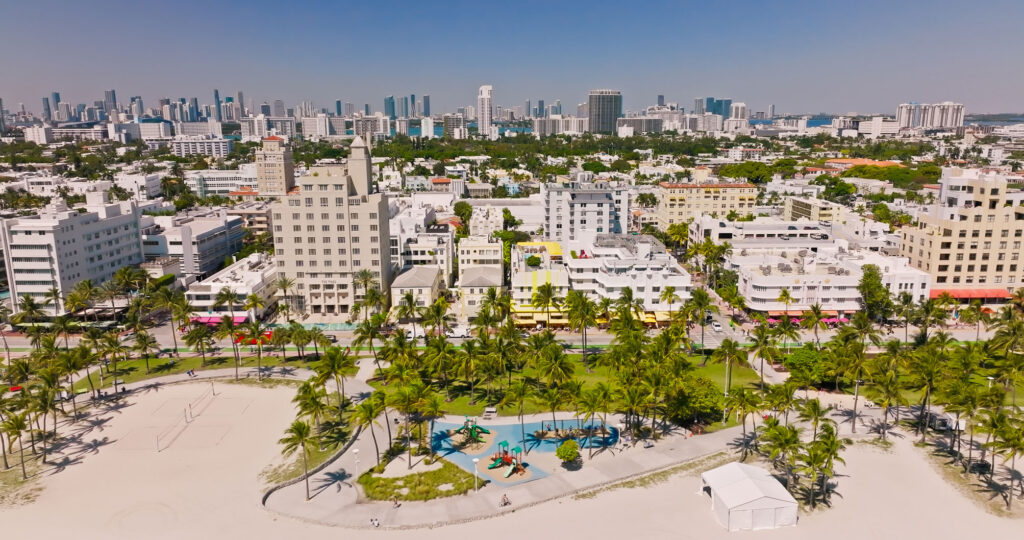 Aerial view of a city beachfront with palm trees, a playground, sandy beach, and high-rise buildings in the background under a clear blue sky.