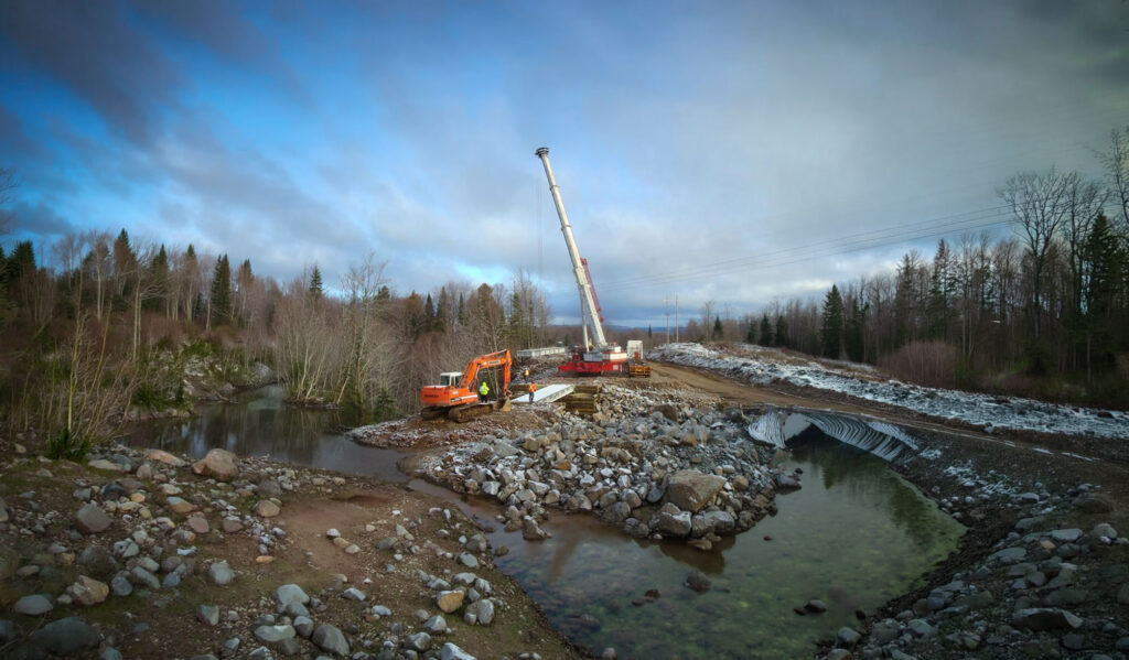 Construction equipment, including an excavator and a crane, operate on a rocky riverbank near a partially cleared area on a cloudy day, with trees and power lines in the background.