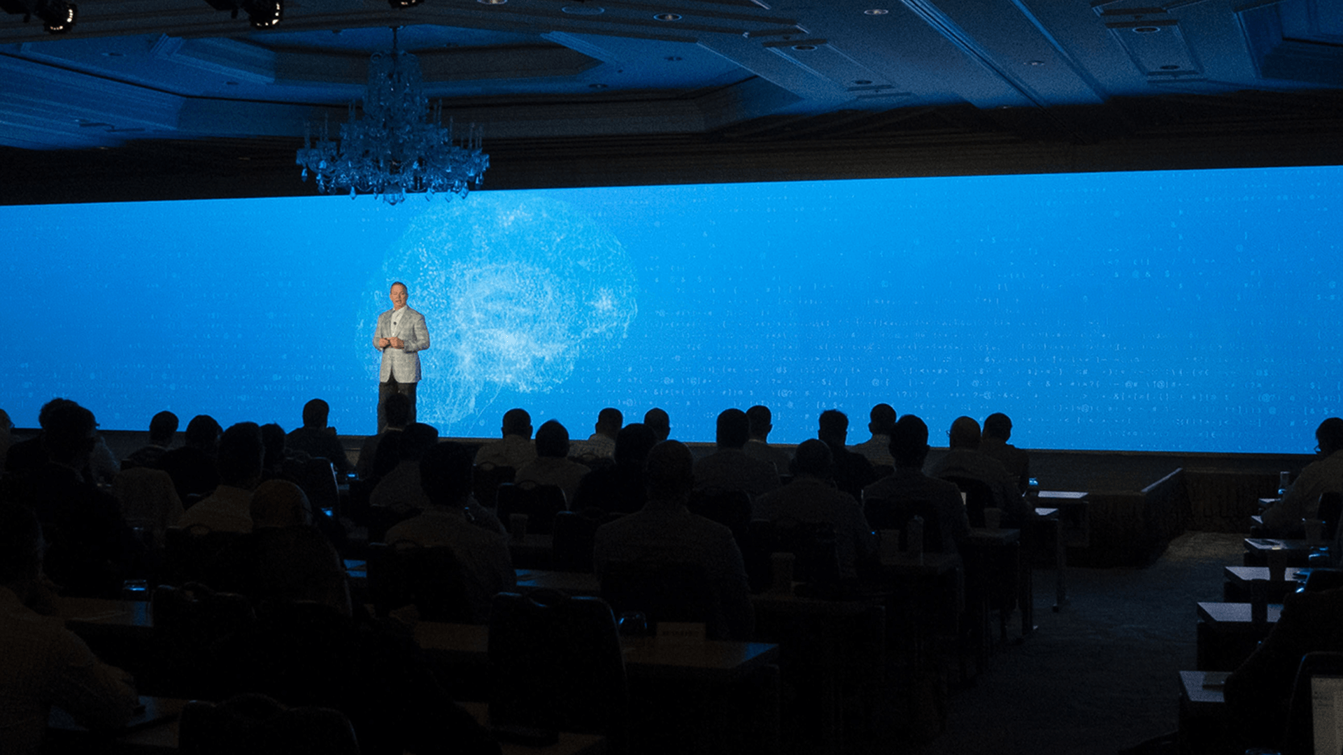 A speaker stands on stage in front of a large blue digital background, addressing an audience seated in a dimly lit conference room.