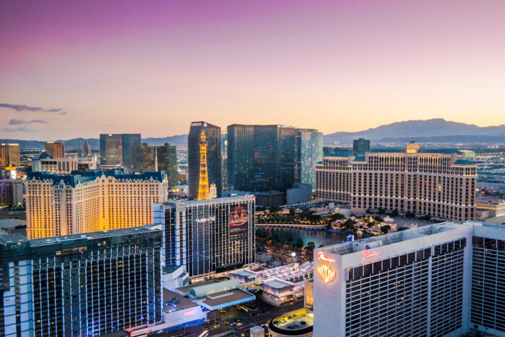 Aerial view of Las Vegas Strip hotels and casinos at sunset, with city buildings, lights, and distant mountains under a purple sky.