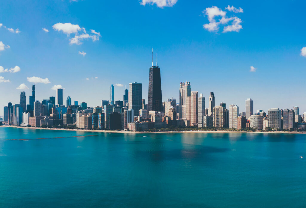 A panoramic view of a city skyline with tall buildings along a lakeshore under a clear blue sky with scattered clouds.