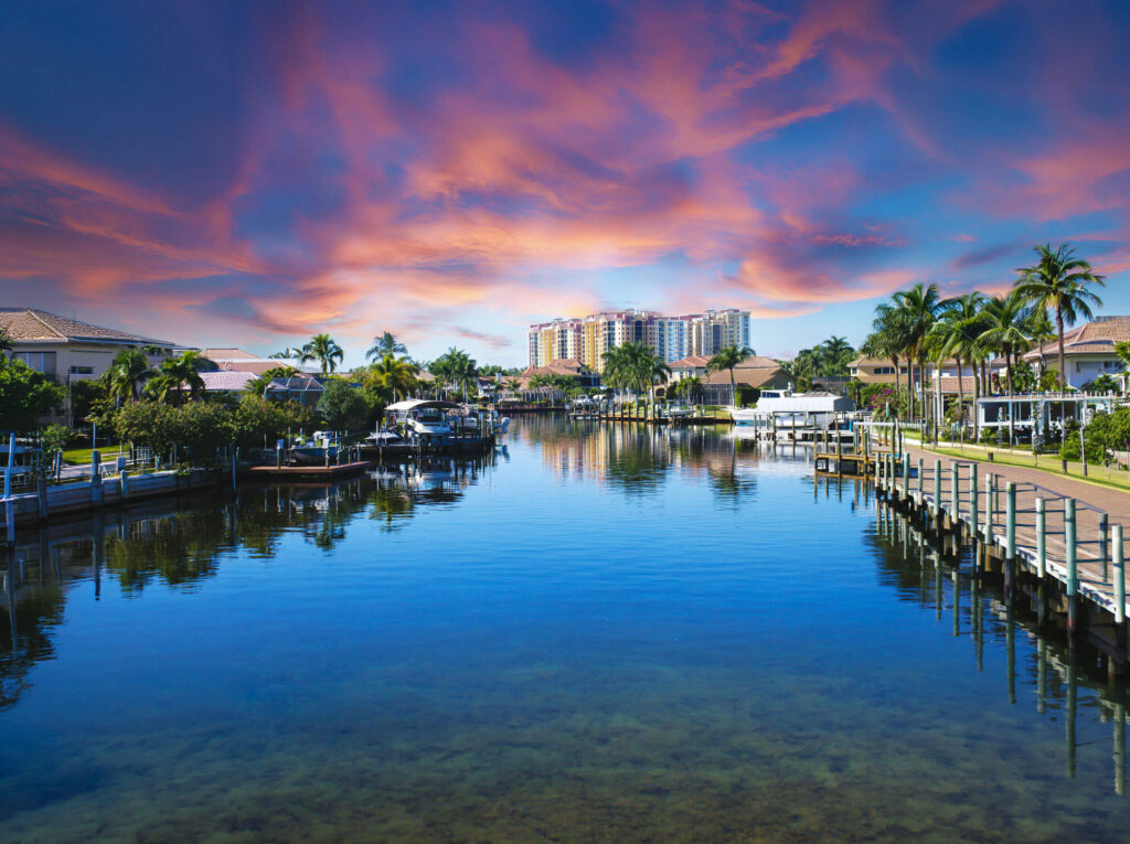 A calm canal lined with docks and palm trees reflects colorful clouds and a pink sunset, with tall buildings in the background.