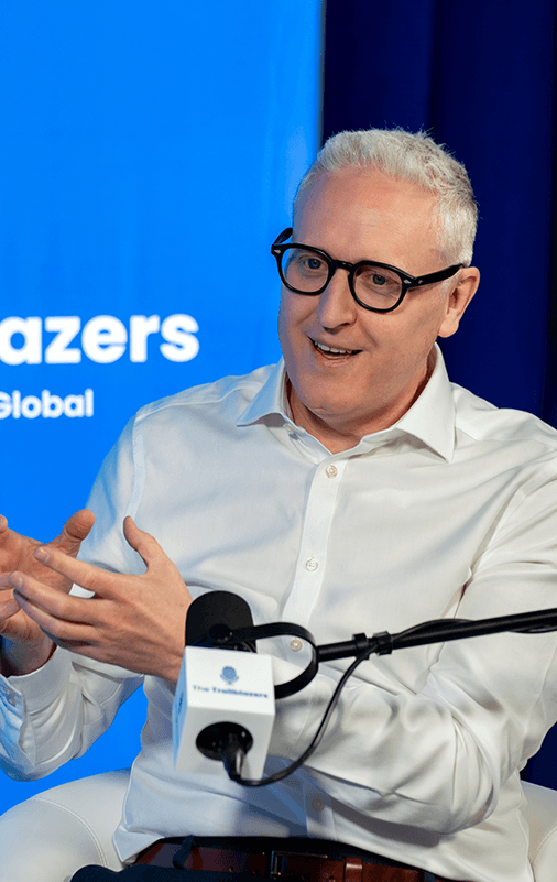 A man with short gray hair and glasses speaks while gesturing with his hands during an interview. A microphone and a blue background with text are visible.