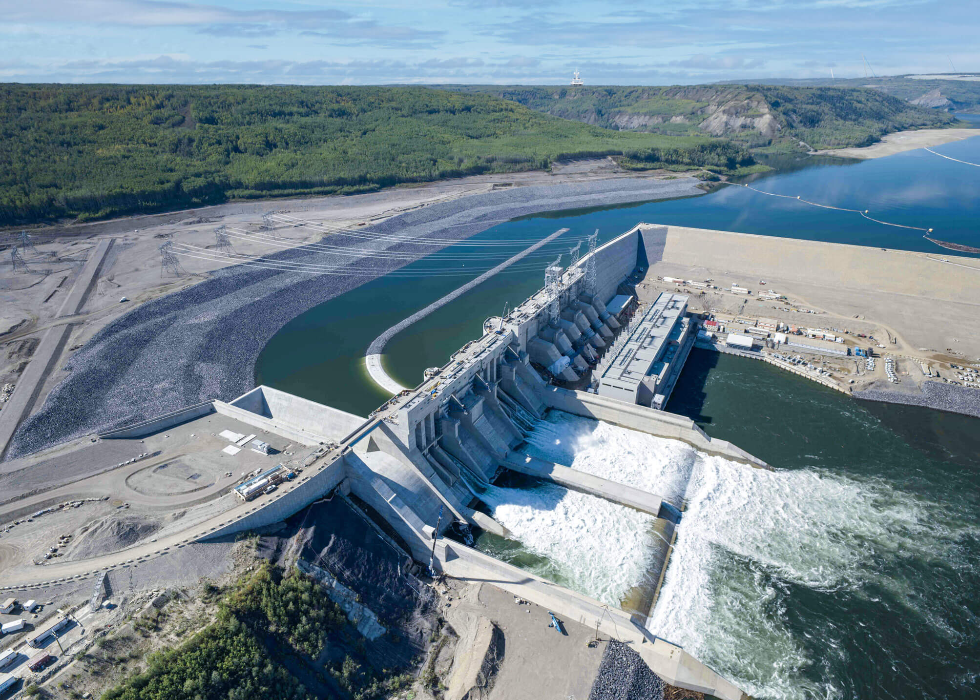 Aerial view of a large hydroelectric dam releasing water, surrounded by forested hills and a river.